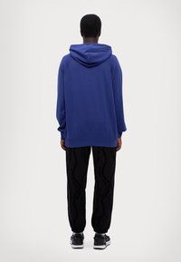 Μπλε φούτερ με κουκούλα και μακριά μανίκια, συνδυασμένο με μαύρο παντελόνι με υφασμάτινο σχέδιο. Φοράει μαύρα αθλητικά παπούτσια.