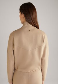 Sudadera beige con un cuello alto, con un detalle de costura en forma de cheurón a lo largo de la espalda y un pequeño logo dorado cerca del cuello.