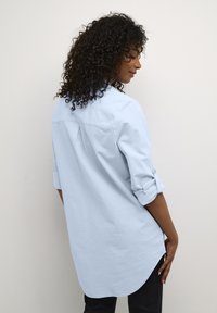 Vaaleansininen pitkähihaista paitaa, jossa on kaartuva helma, napilliset kalvosimet ja takana laskosdetaili, valmistettu pehmeästä, teksturoidusta kankaasta.