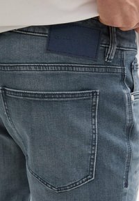 Sötétkék farmernadrág, kopott textúrával, bőr övszíjjal a hátsó derékrészen és dupla varrású hátsó zsebekkel.