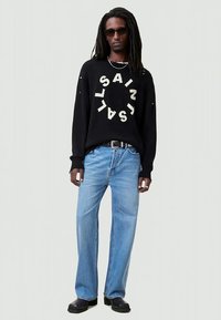 Homem com dreadlocks longas e óculos de sol, veste um suéter preto com texto circular branco, jeans azul claro, sapatos pretos e um colar de corrente prateada.