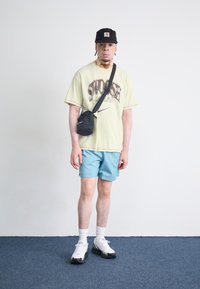 T-shirt gráfica amarela com o texto "Swoosh", calções azul claro, mochila preta, meias brancas e ténis brancos/pretos com detalhes verdes.