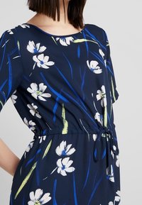 Laivastonsininen kukkamekko, joka on valmistettu kevyestä kankaasta ja jossa on valkoisia kukkia ja vihreitä yksityiskohtia. Mekossa on nyöri vyötäröllä, joka mahdollistaa säädettävän istuvuuden.