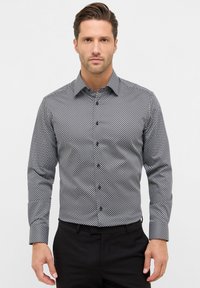 Homme portant une chemise à motifs géométriques noirs et blancs et un pantalon noir, se tenant contre un fond clair uni.