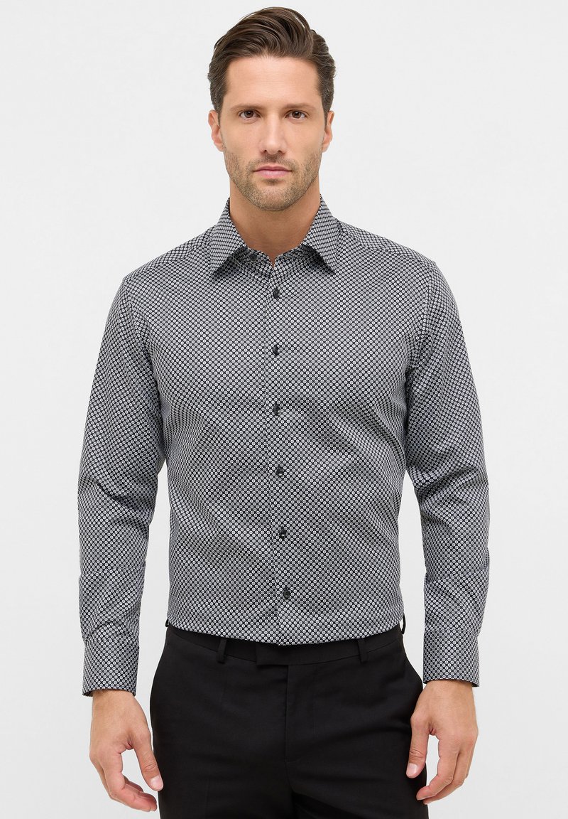 Homme portant une chemise à motifs géométriques noirs et blancs et un pantalon noir, se tenant contre un fond clair uni.