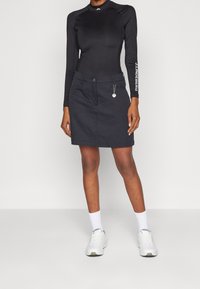 Žena oblečená v černém dlouhém rukávu a černé sukni s jemným stříbrným řetízkovým detailem, s bílými ponožkami a bílými teniskami, stojící před jednoduchým pozadím.