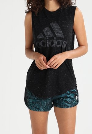 Mujer con camiseta negra sin mangas de Adidas y pantalones cortos deportivos verdes oscuros con estampado, de pie con las manos entrelazadas.