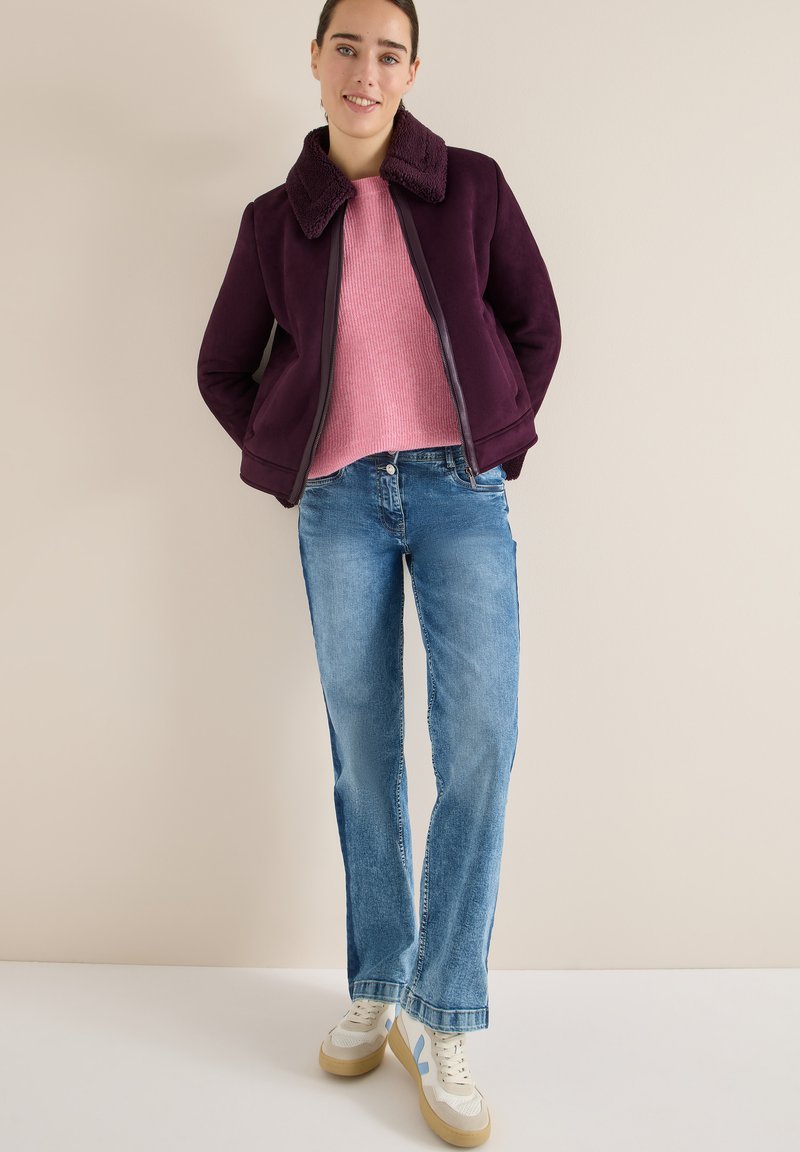 Giacca in finta suede bordeaux con colletto testurizzato, maglione rosa a coste, jeans azzurri chiari e sneakers crema con accenti blu.