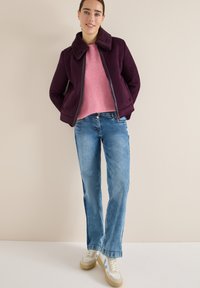 Giacca in finta suede bordeaux con colletto testurizzato, maglione rosa a coste, jeans azzurri chiari e sneakers crema con accenti blu.