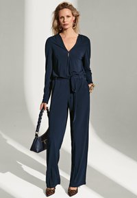 Navyblauw jumpsuit met een diepe V-hals, lange mouwen, een strikceintuur in de taille en een wijde pijpdesign. Accessoires omvatten een gegraveerde handtas en hakken.
