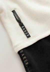 Sweatshirt présentant une partie supérieure crème et une partie inférieure noire, avec une fermeture éclair sur le côté et une étiquette affichant "BAKER" en blanc sur fond noir.