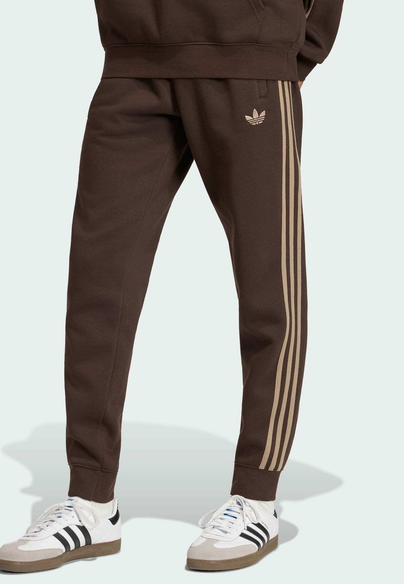 Personne portant un pantalon de survêtement Adidas marron avec des rayures et un logo beige, associé à des baskets Adidas blanches et noires, debout sur un fond clair.