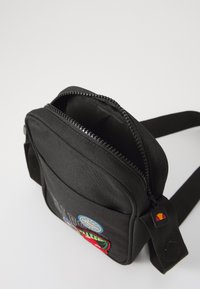 Borsa a tracolla nera in tessuto con chiusura zip, tasca frontale con toppe colorate, tracolla regolabile e superficie texturizzata.