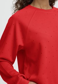 Vrouw gekleed in een rode lange mouw top versierd met kleine rode kralen verspreid over de stof, hand op haar heup rustend.