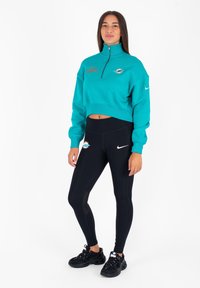 Felpa con zip turchese con logo ricamato, abbinata a leggings neri a vita alta con logo. Le scarpe sportive nere completano l'outfit.