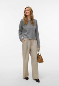 Hall hallisvalge äärisega kampsun, beežid volditud püksid ja pruun käekott. Modellil on pikad juuksed ning ta seisab vabalt, üks käsi taskus.