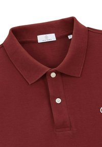 Polo shirt bordeaux avec un col et deux boutons blancs, présentant un petit logo partiellement visible sur le côté droit.