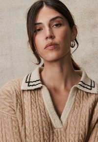 Beige gebreide trui met kabelpatroon, voorzien van een lichtgekleurde kraag met zwarte strepen. Model heeft middellang donker haar en oorbellen met een ring.
