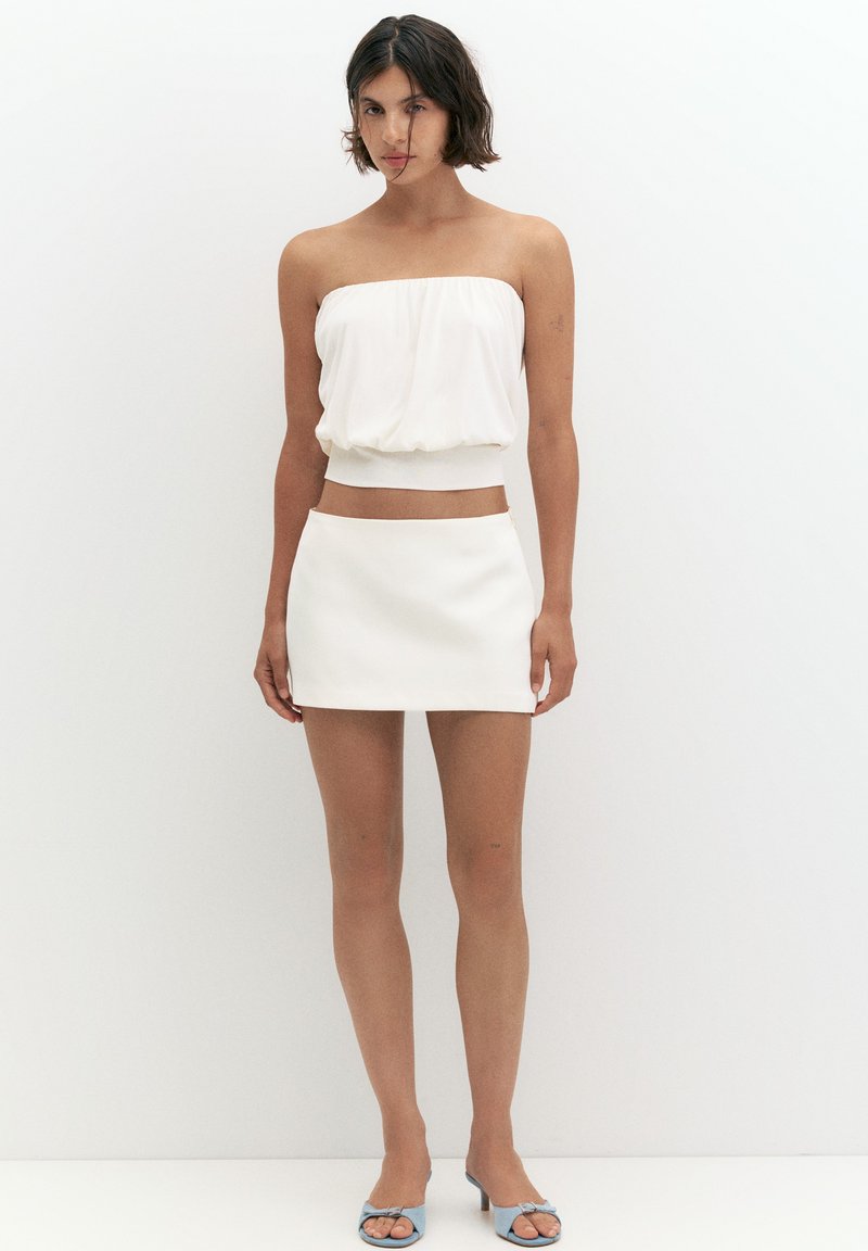 Femme aux cheveux courts et foncés portant un top blanc sans bretelles, une mini-jupe blanche et des talons ouverts bleu clair, debout contre un mur blanc uni.
