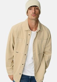 Man in een beige ribfluwelen jas over een wit shirt, blauwe spijkerbroek en een crèmekleurige gebreide muts, poserend tegen een effen achtergrond.