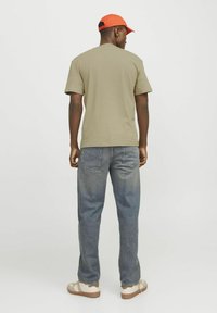Khaki T-shirt, korte mouwen, comfortabele pasvorm, gecombineerd met blauwe spijkerbroek. Witte sandalen met beige en bruin accent. Rode pet op het hoofd.