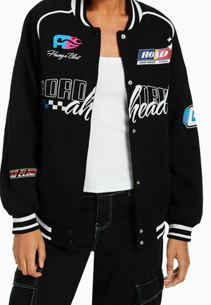 Veste varsity noire avec des bandes blanches aux poignets et au col, patchs de course colorés, portée sur un top blanc et un pantalon noir avec coutures blanches.