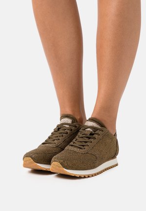 Personne portant des baskets à lacets texturées vert olive avec des semelles intermédiaires blanches et des semelles extérieures en gomme naturelle, debout sur une surface blanche.