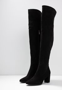 Kaksi korkeaa mustasta mokasta valmistettua saapasta, joissa on paksut korot ja sivuvetoketjut, seisoo pystyssä valkoisella pinnalla valkoista taustaa vasten.