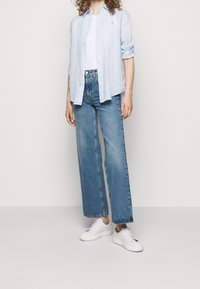 Person som bär en ljusblå knappad skjorta över en vit topp, blå raka jeans och vita snörskor, stående mot en enhetlig bakgrund.