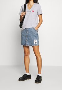 Šedé tričko s V-výstřihem a červeným logem "Calvin Klein", spárované s džínsovou sukní s předními knoflíky a bílou nášivkou CK. Černé boty.