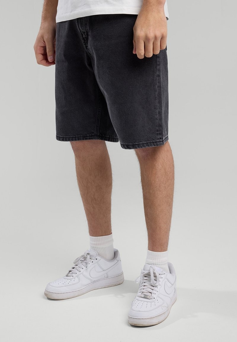 Mies, jolla on päällään mustat polvimittaiset shortsit, valkoiset perusnilkkasukat ja valkoiset Nike-tennarit, seisoo vaaleanharmaata taustaa vasten.