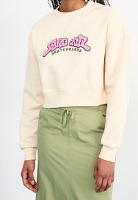 Felpa cropped color crema realizzata in tessuto morbido, con un logo grafico rosa e nero; indossata con una gonna verde chiaro con coulisse.