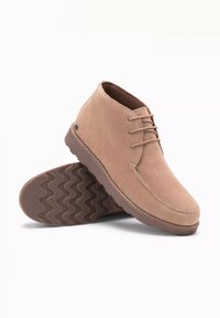 Botas de tobillo de ante beige con punta redonda y cordones, que presentan una suela de goma marrón texturizada con un patrón de zigzag.