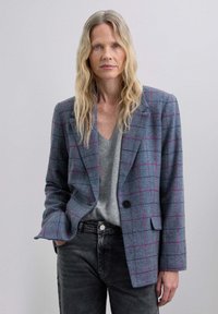 Blazer de cuadros gris con acentos rosas, con solapa de muesca y cierre de un solo botón, llevado sobre un suéter gris de cuello en V y jeans negros.