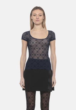 Donna che indossa una maglia trasparente blu navy con motivi e maniche corte, una minigonna nera e calze con motivi abbinati, in piedi di fronte alla fotocamera.