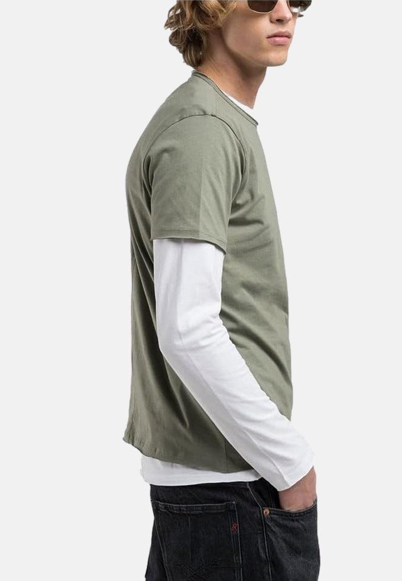 Un T-shirt vert olive à manches courtes par-dessus une chemise à manches longues blanche, avec une coupe décontractée et un col rond. Porté avec un jean sombre.