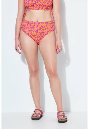 Frau trägt pink-orange florale Bikinihose mit hohem Bund und passendem Oberteil, dazu pinke Sandalen und Ringe an den Fingern, steht vor einer weißen Wand.