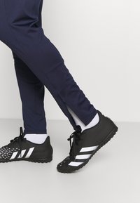 Musta urheilukengät, joissa on valkoisia raitoja, teksturoitu päällinen ja kumipohja. Kengät on yhdistetty tummansinisiin urheiluhousuihin, joissa on sivuvetoketjut.