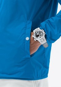 Lyseblå, vandtæt jakke med knapdetalje. Et hvidt digitalt ur med et mønstret urskive er synligt på en hånd i jakkens lomme.