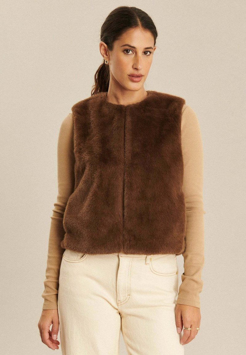 Brązowa kamizelka z futra ekologicznego z okrągłym dekoltem, o gładkiej fakturze i zapięciu z przodu, noszona na beżowej bluzie z długim rękawem oraz jasnych dżinsach.
