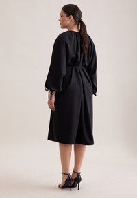 Czarna sukienka z długim rękawem, z taliowaniem i rozkloszowanym dołem; posiada wysoki kołnierz oraz marszczenia na rękawach i w talii.