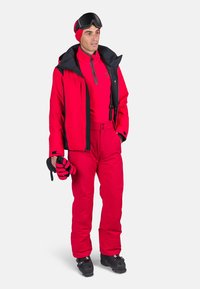 Red skiing outfit consisting of a jacket and pants. Features a zip-up jacket, gloves with black accents, and black ski goggles.