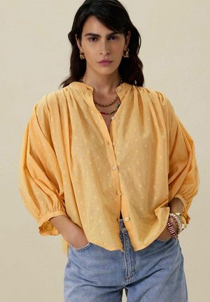 Chemise jaune à pois, boutonnée avec un col mandarin et des manches bouffantes surdimensionnées, assortie à un jean bleu clair et des bracelets en couches.