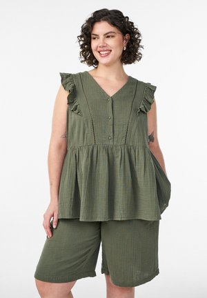 Lachende vrouw met krullend haar, gekleed in een mouwloze olijfgroene top met knopen en ruches, gecombineerd met bijpassende knielange shorts, met één hand in de zak.