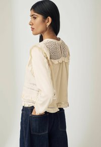 Kvinde med langt sort hår iført en cremet bluse med blondelæg og flæser, hænderne i lommerne på mørke denimjeans, der kigger til venstre i en neutral indstilling.