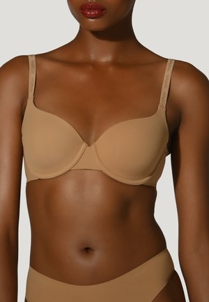 Kvinde iført en sømløs beige BH og matchende undertøj, med mærkede stropper og blank rød læbestift, mod en ensfarvet baggrund.