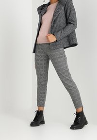 Harmaa villasekoitejakku, jossa epäsymmetrinen vetoketju, ja pehmeä lila neule, yhdistettynä ruudullisiin kapeneviin housuihin ja mustiin nauhallisiin saappaisiin.