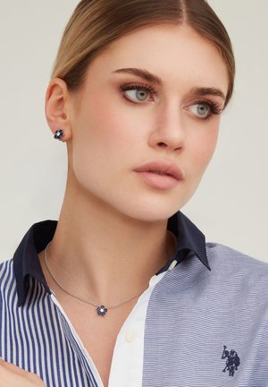 Donna dalla pelle chiara che indossa orecchini e collana abbinati a forma di fiore blu, vestita con una camicia a righe blu e bianca con colletto.