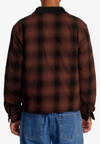 Brun och svart rutigt flanneljacka med en kordfloderkant, som har en avslappnad passform och knäppta manschetter. Synlig sömnad och texturer.