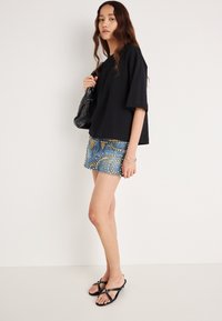 Čierne nadrozmerné tričko, modré riflové šortky so svetlými zlatými kruhovými cvočkami a čierne žabky. Taška je vyrobená z lesklého materiálu.